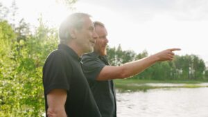 Två män står riktade till höger i bild, och står framför en sjö och skog en solig dag. En av männen pekar med ett finger för att visa vad han tittar på.